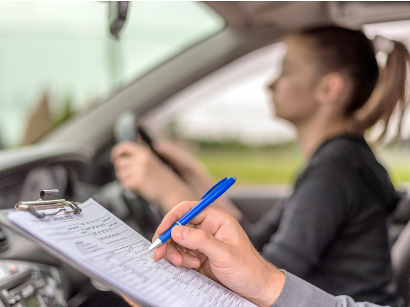 Person with ponytail driving out of focus with focus on a test on a clipboard with a hand holding a pen