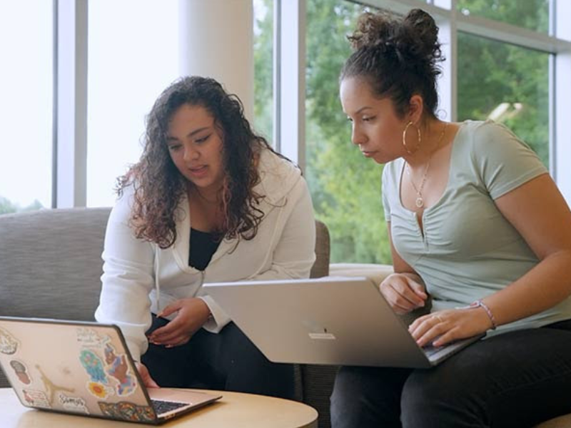 Two women of Hispanic descent holding laptops sitting and looking at one screen.