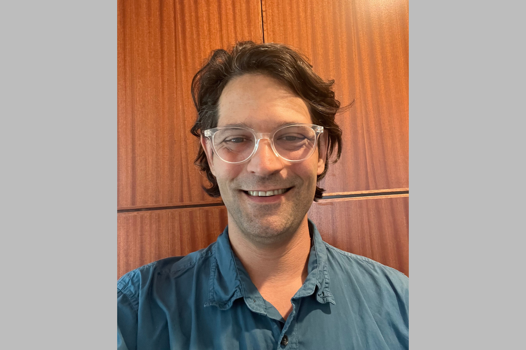 Haas A man with brown, medium length hair, a blue button up shirt, and clear-framed classes standing in front of a woodpaneled wall.