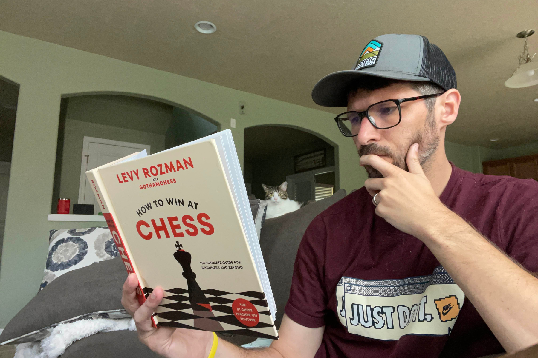 Chris Mock Chris Mock sitting on a couch in a living room wearing a grey hat and maroon shirt that says "Just Do It." He is wearing glasses and reading a book with the text "Levy Rozman aka Gothamchess How to Win At Chess" on the cover