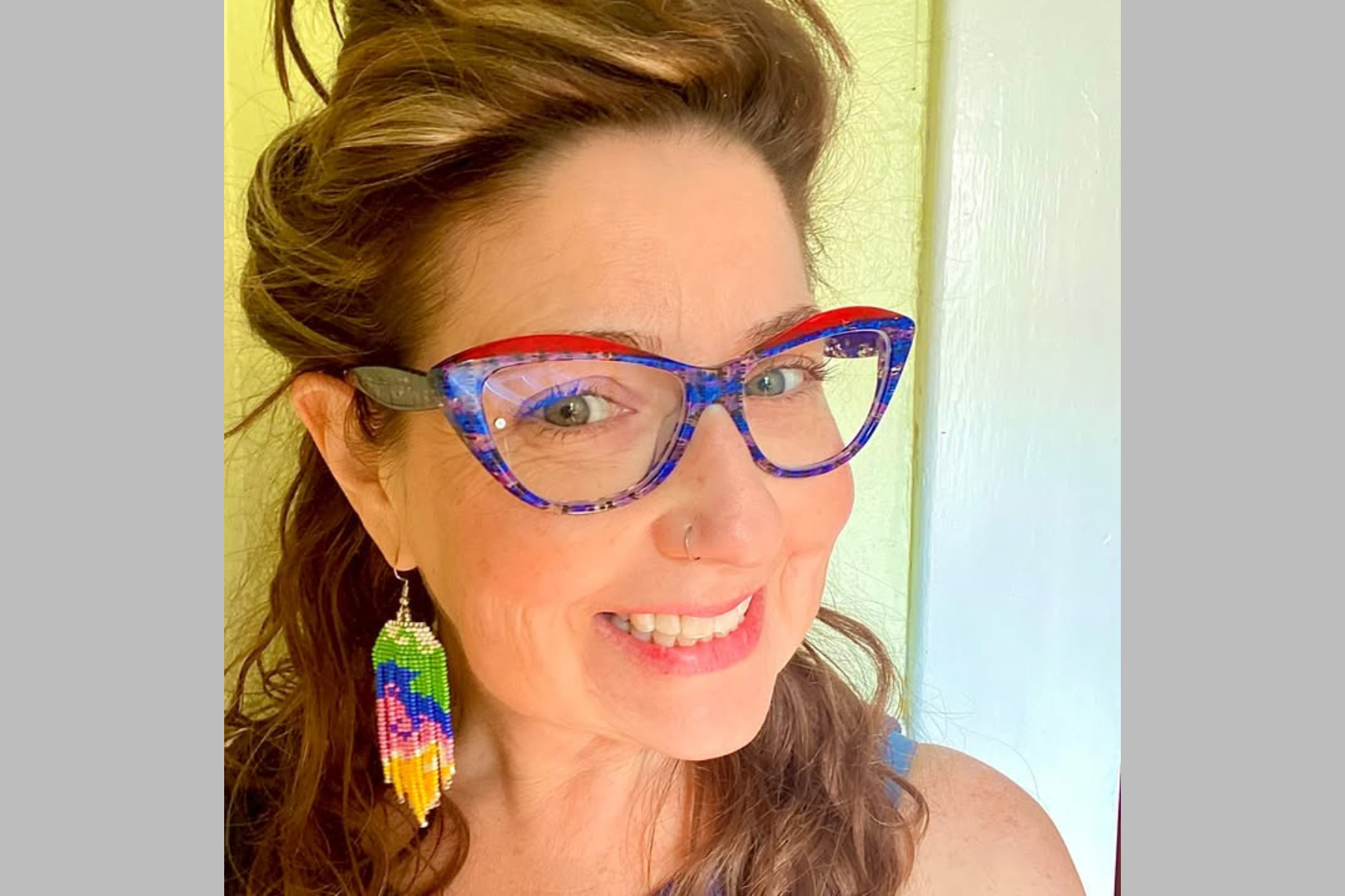 Tandy Tillinghast Tandy Tillinghas smiling wearing red, blue, and purple glasses and colorful dangling earrings.