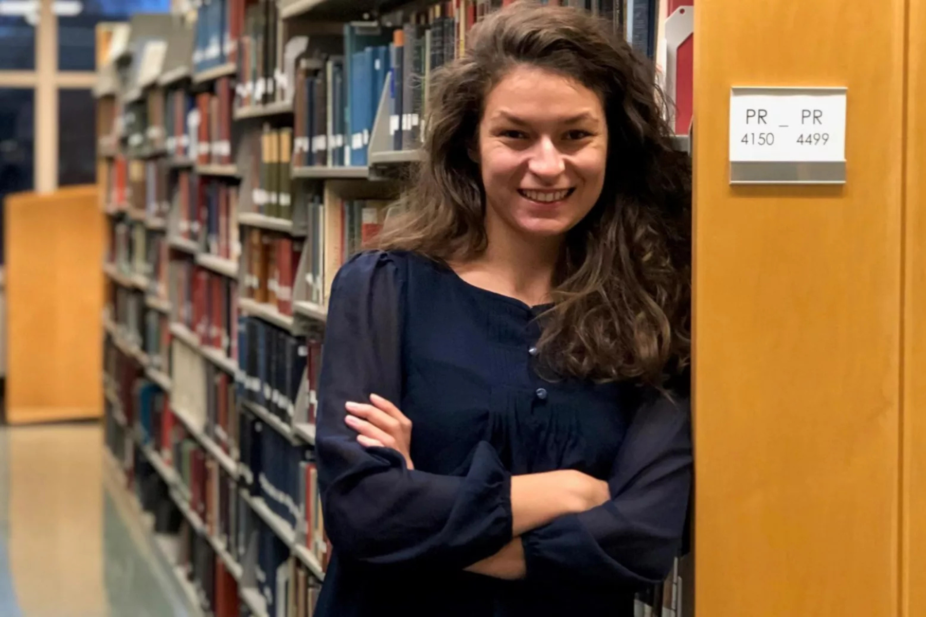 Vanessa Petroj Vanesssa Petroj, a woman with a dark blue blouse, smiling and leaning against a library bookshelf. Her arms are crossed and she has voluminous brown hair that goes past her shoulders.