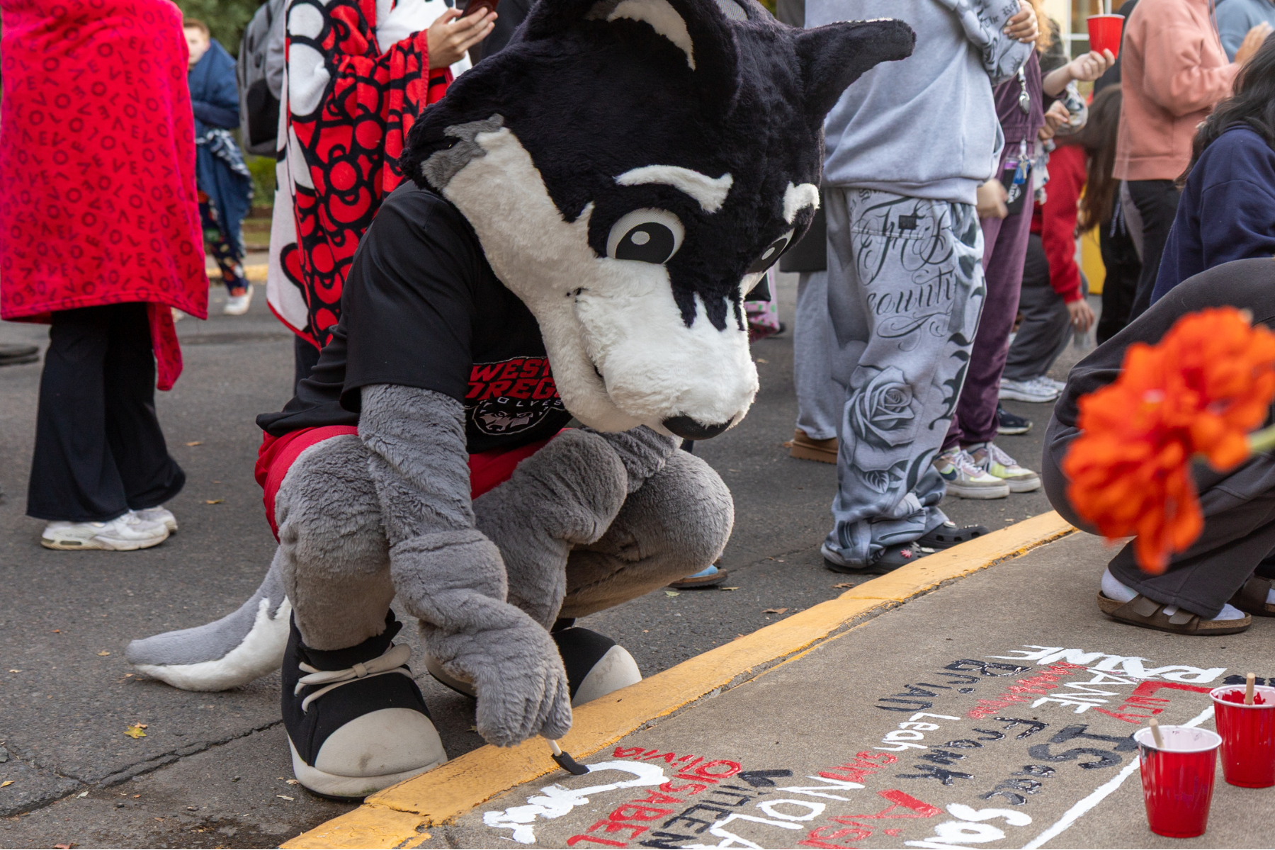 WOU Mascot, Wolfie, crouching towards the ground with a paint brush, painting his name in white on a sidewalk with students surrounding them.