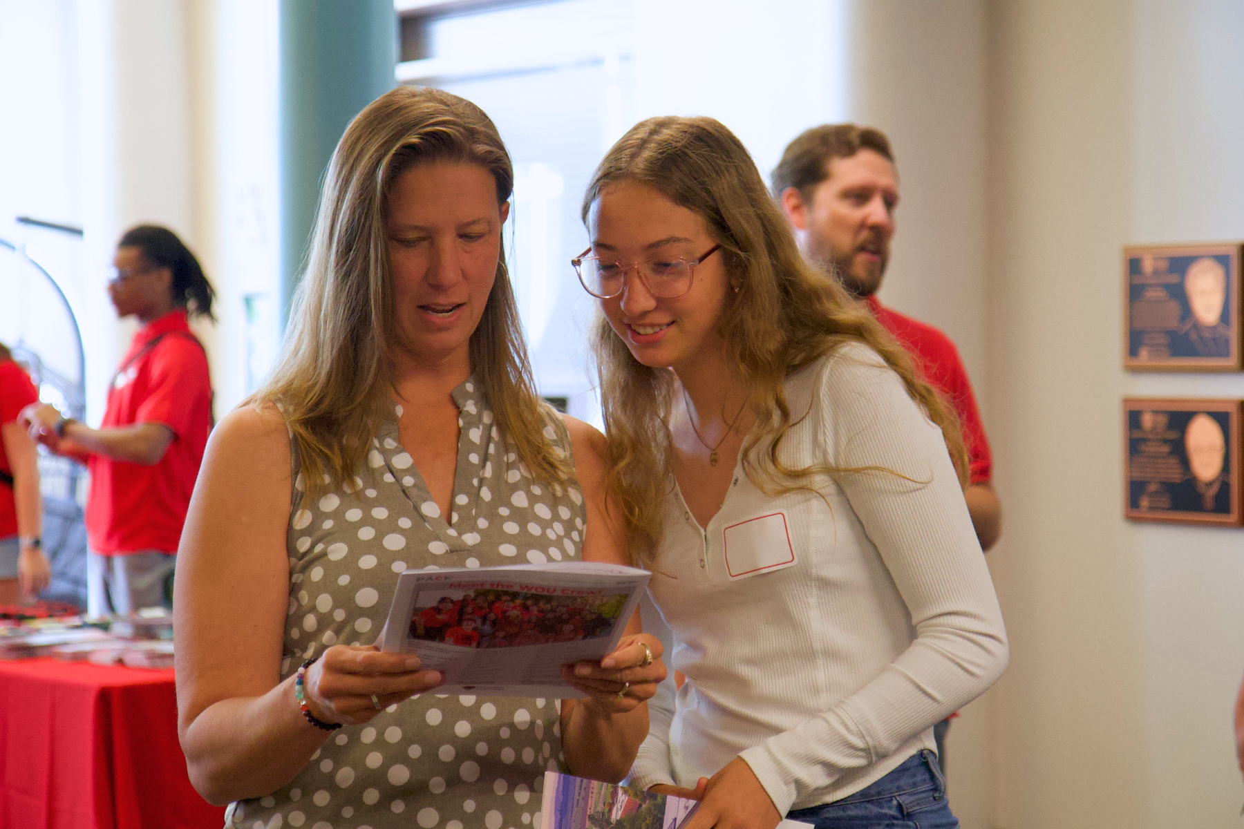 Two women standing next to each other reviewing a piece of paper. The woman on the left is wearing a taupe sleeveless top with white polka dots. The woman on the right is wearing a long sleeve white shirt and blank nametag.