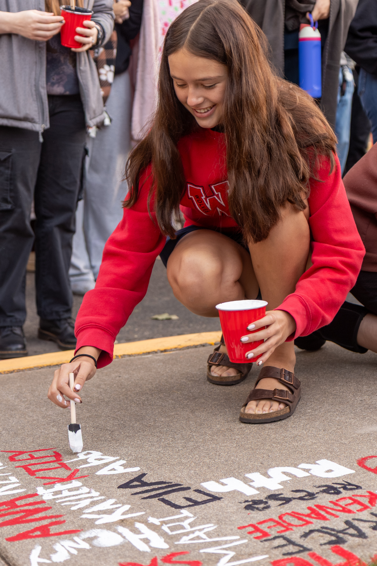 A woman with long brown hair, a red WOU Wolves sweater, shorts, and brown sandals paints her name on the sidewalk  with one hand with a red cup of pain in the other hand.