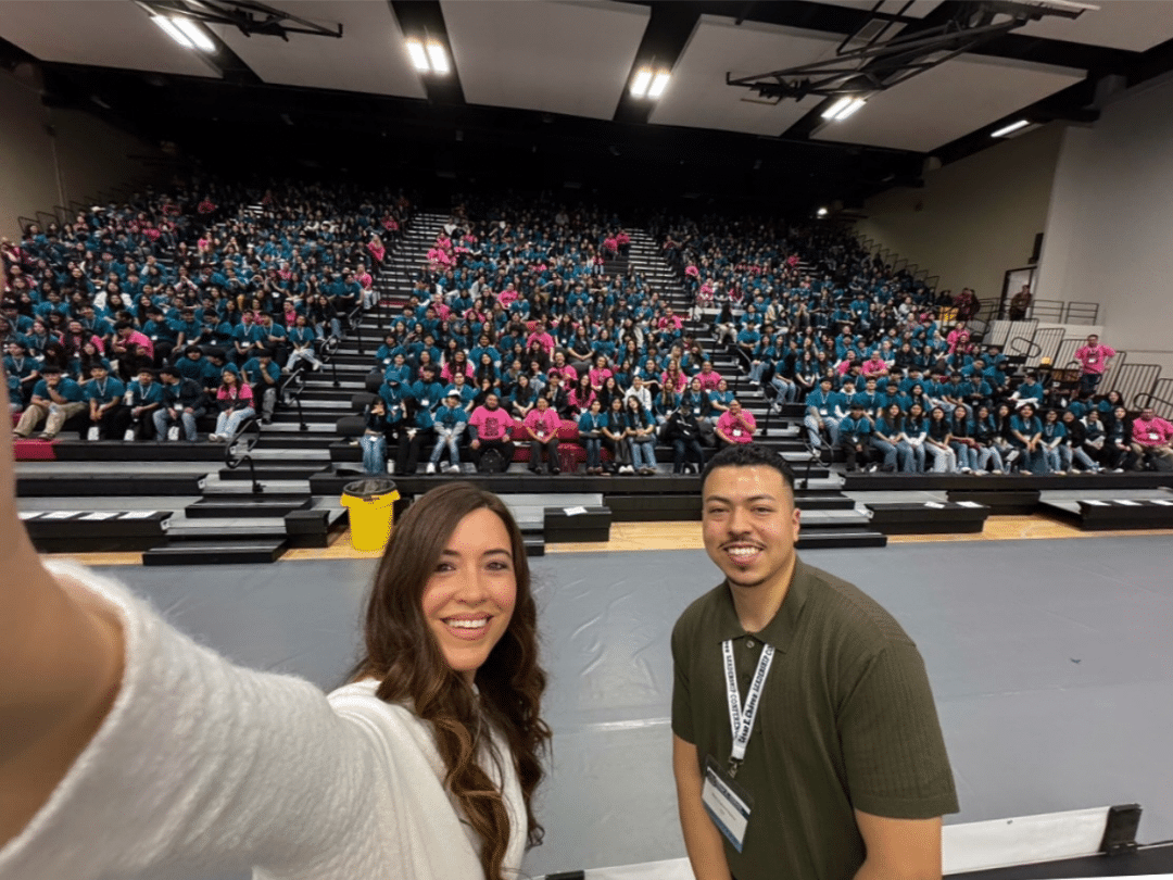 CECLC PR Two smiling individuals pose for a selfie at the 2025 César E. Chávez Leadership Conference. Behind them, a packed auditorium with attendees wearing teal and pink shirts fills the elevated bleacher seating, indicating a large and engaged audience at the event.