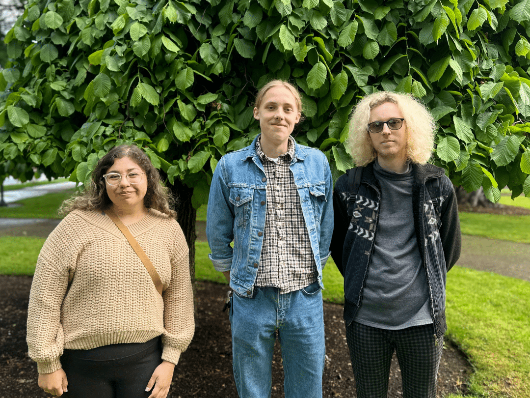 Peter Sears PR Three young adults stand side-by-side outdoors in front of a leafy green tree. From left to right: A person with shoulder-length dark hair and glasses wears a beige v-neck sweater. The person in the middle has light brown, shoulder-length hair and is wearing a blue denim jacket over a plaid shirt and jeans. The person on the right has curly, light blonde hair, sunglasses, a dark grey jacket with a geometric pattern, and plaid pants.