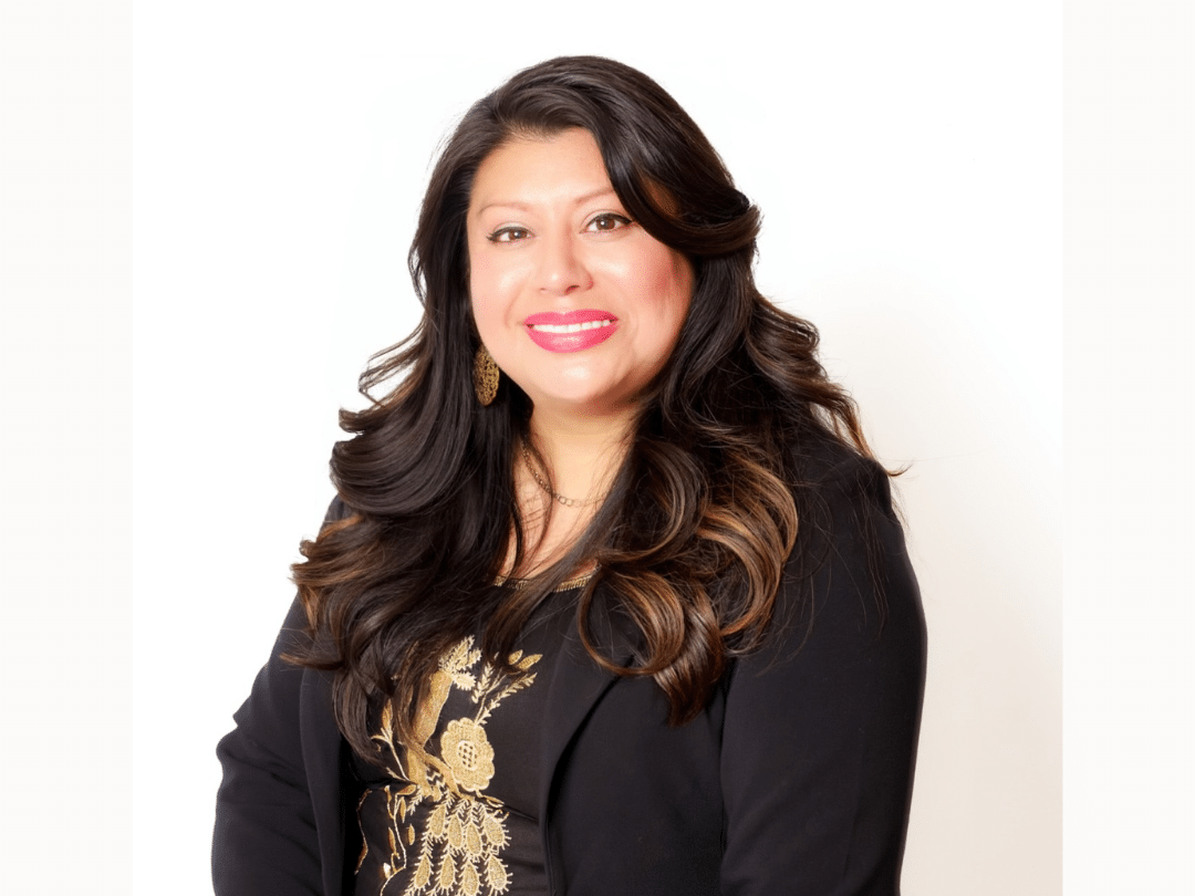 Press Release Photo Size (3) A smiling woman with long, dark, wavy hair, wearing a black blazer over a dark top with gold embellishments, and gold hoop earrings, poses against a plain white background.