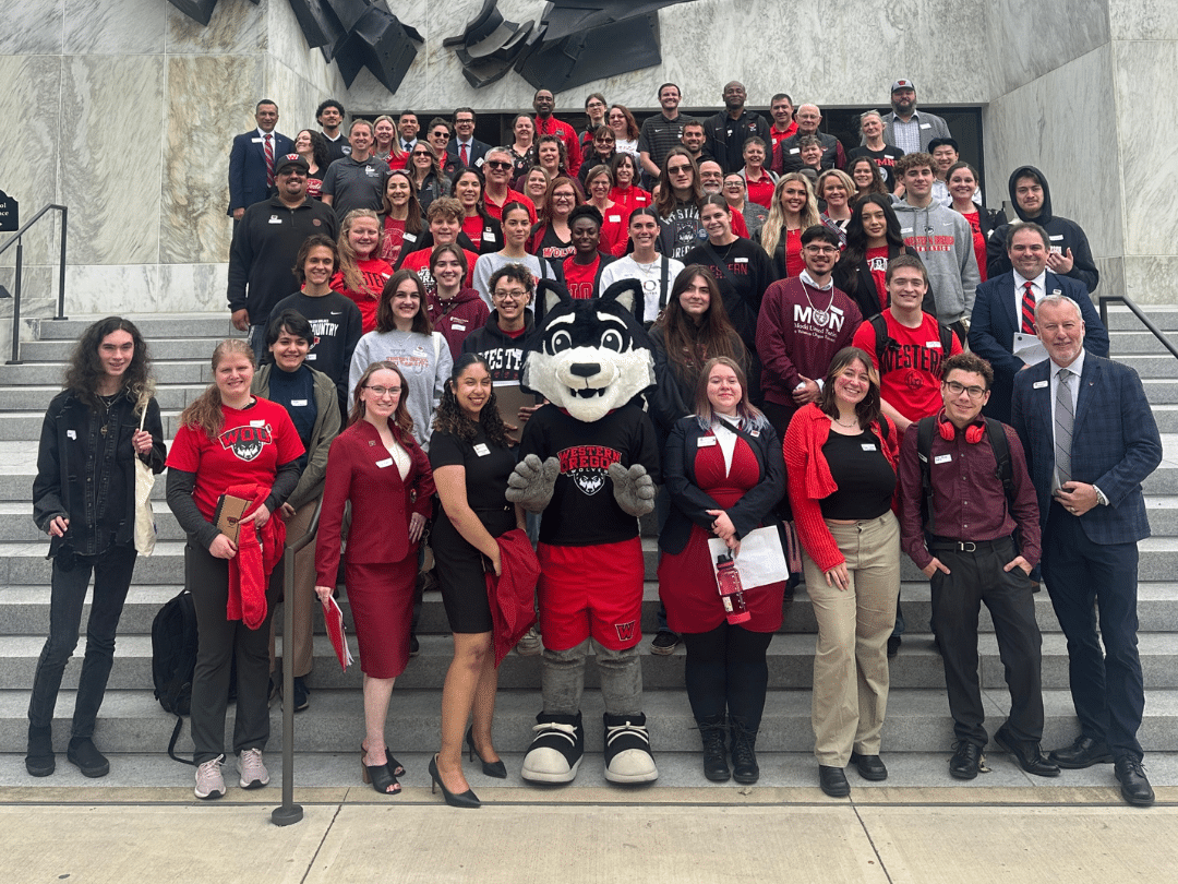 A large group of students and adults, many wearing red shirts or jackets, pose for a photo on a grand staircase. In the center stands a person in a wolf mascot costume, also dressed in red and black. The group appears to be associated with an educational institution, possibly Western Oregon University, given the "WESTERN OREGON" text visible on some shirts and the mascot's appearance. The setting looks like a public building, possibly a state capitol or courthouse, with classical architecture in the background.