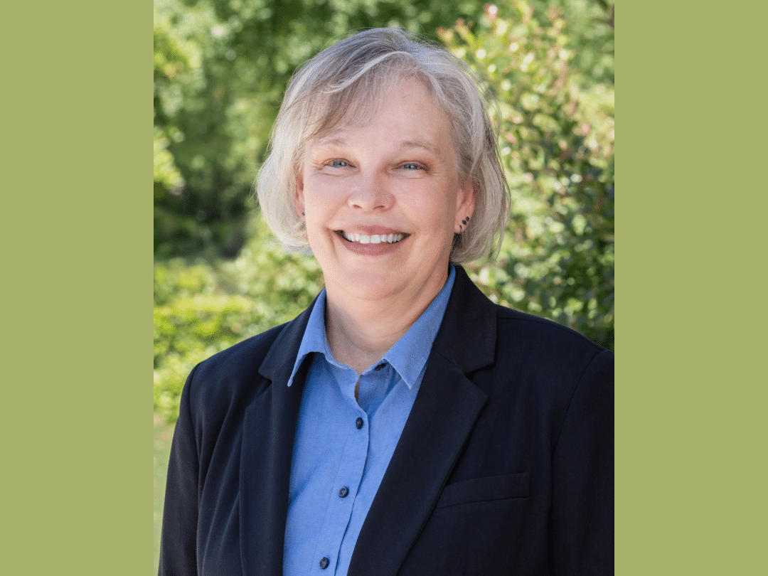 Press Release Photo Size (6) A headshot of a smiling woman with short, gray hair and fair skin. She is wearing a black blazer over a light blue collared shirt. She has small earrings and appears to be outdoors with green foliage in the blurred background.